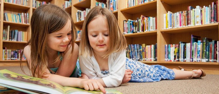 Zwei Mädchen liegen am Boden und lesen in einem Buch. Im Hintergrund befinden sich Bücherregale