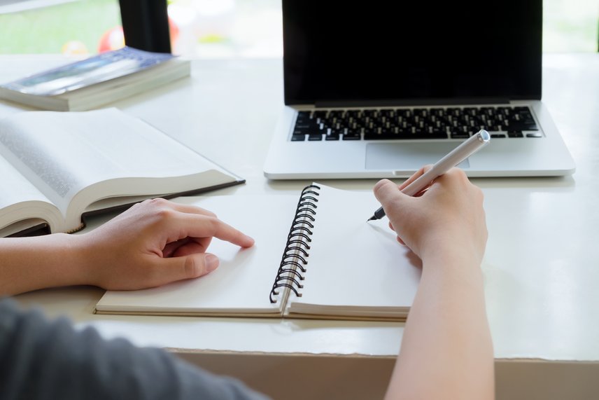 Mensch sitzt bei Tisch schreibt im Heft mit Laptop im Hintergrund